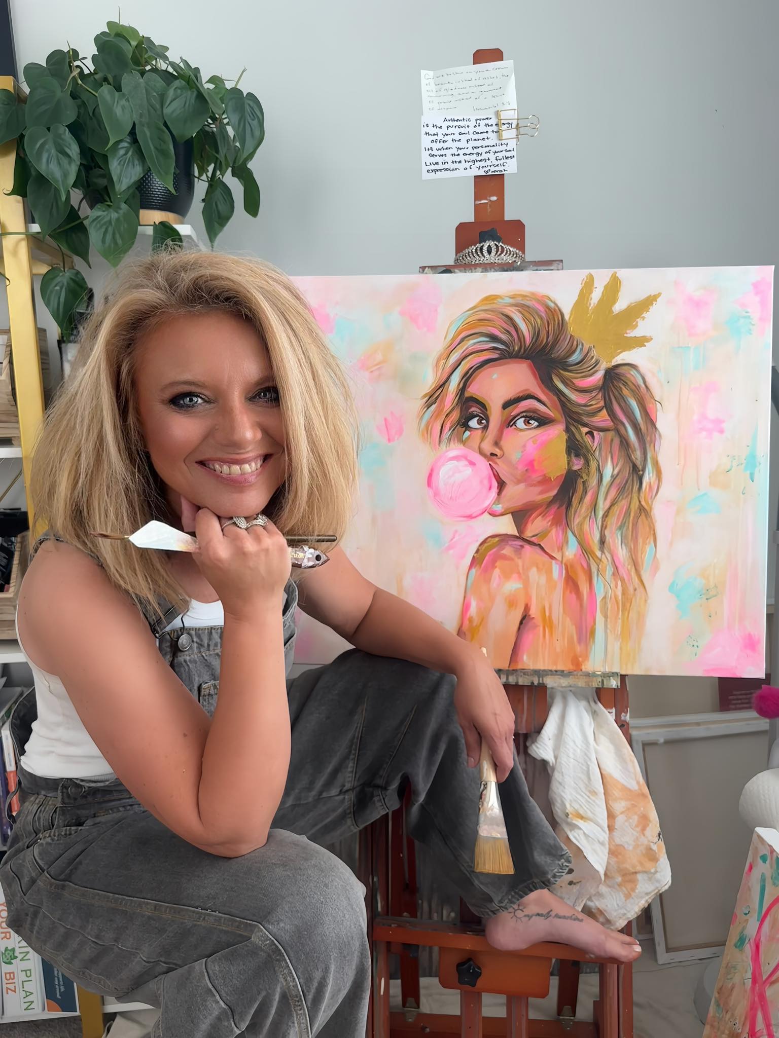 Woman sitting next to a colorful painting of a woman with a crown and pink bubble gum, with a plant and easel in the background.