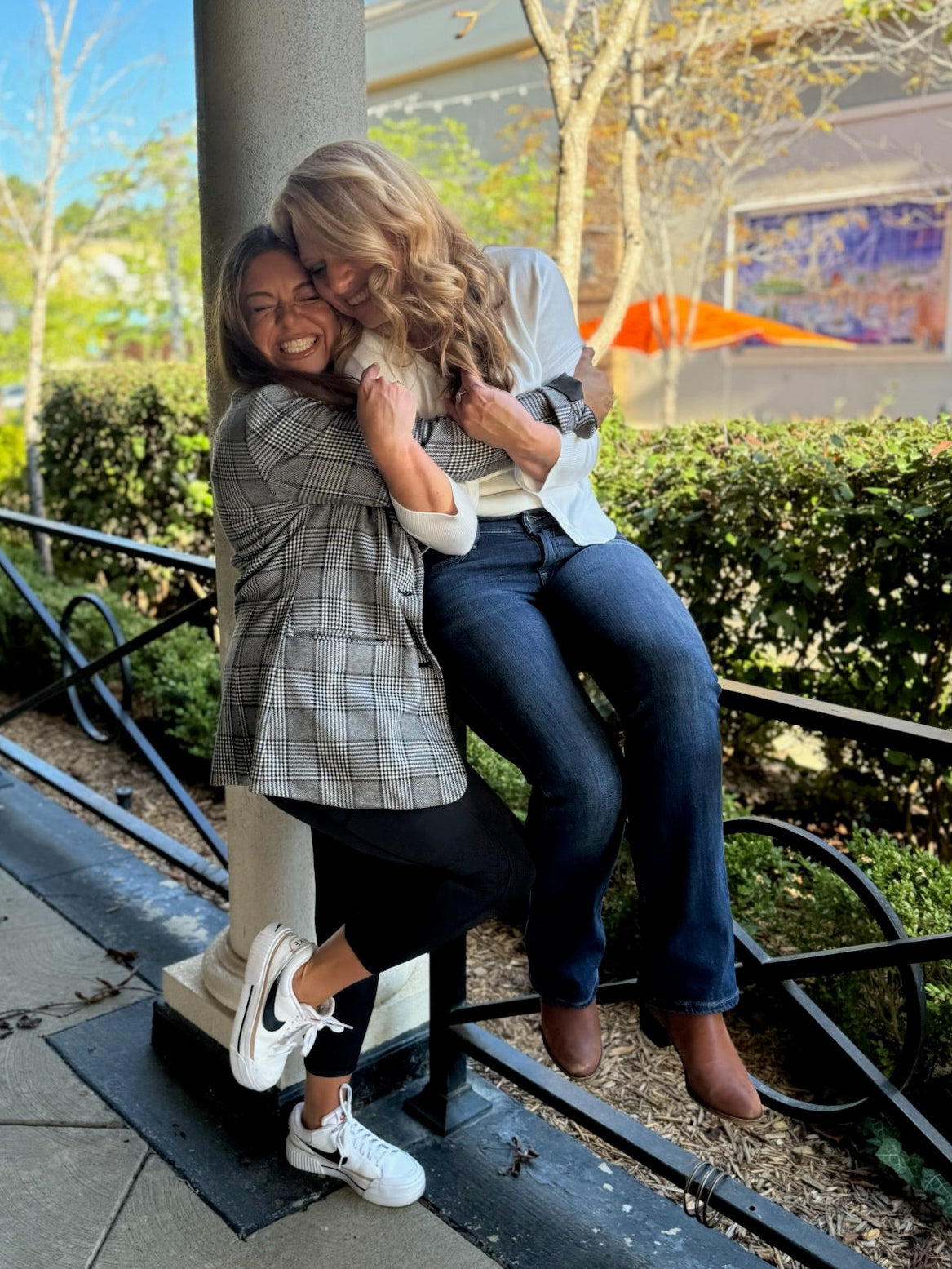 Two women hugging on a sidewalk with a building and trees in the background